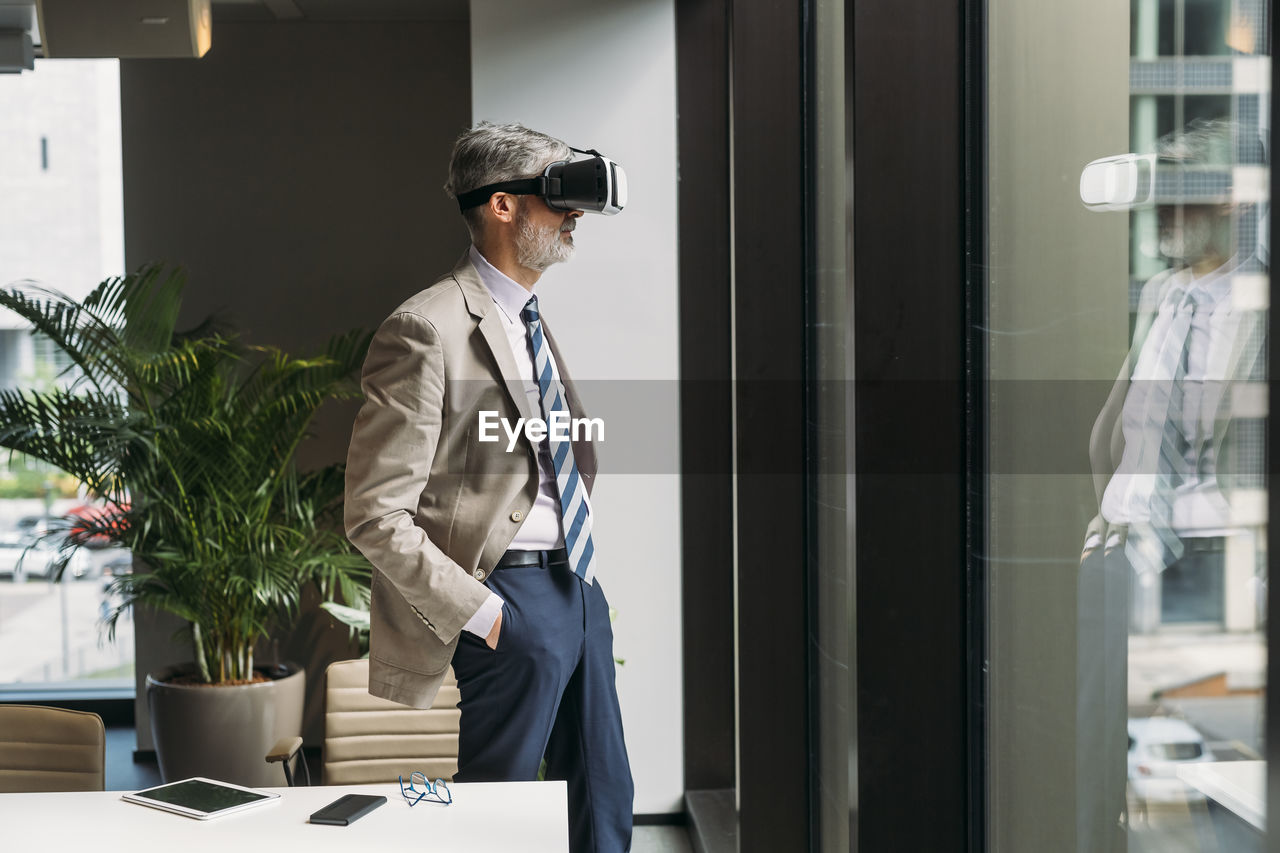 Businessman wearing virtual reality simulator standing with hands in pockets by window in office