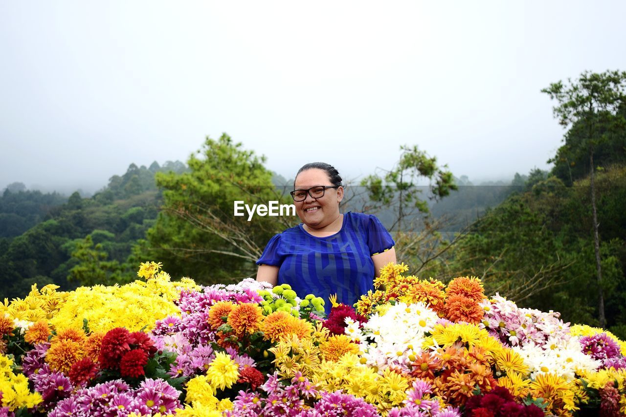 Portrait of smiling woman with various flowers against sky