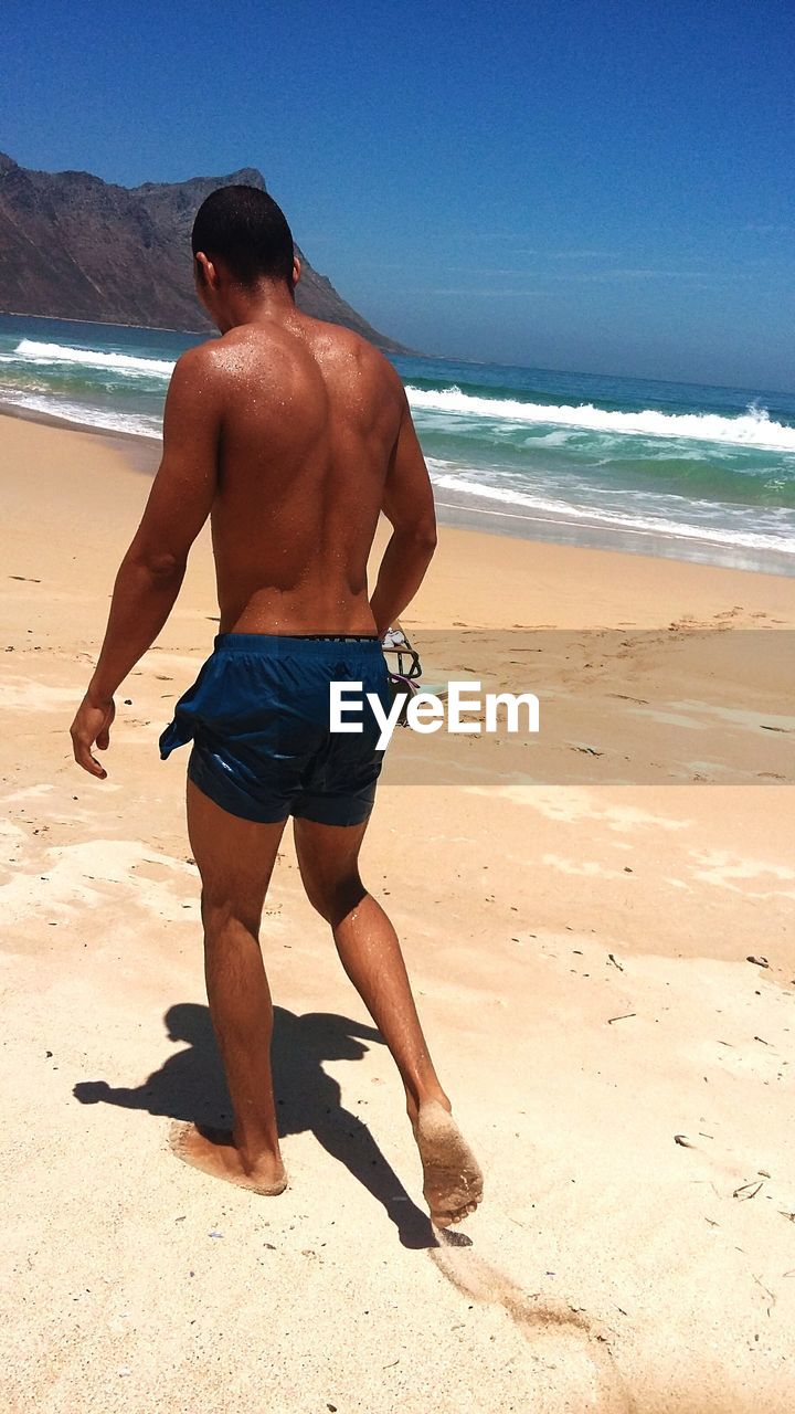 Rear view of shirtless man walking at beach against clear sky