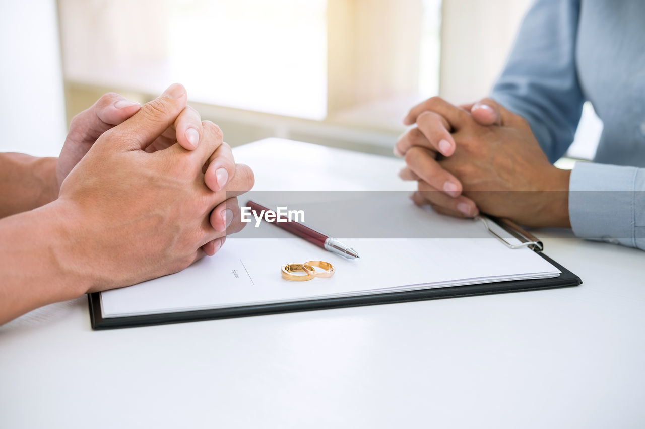 Midsection of couple sitting with divorce paper and rings on desk in courtroom