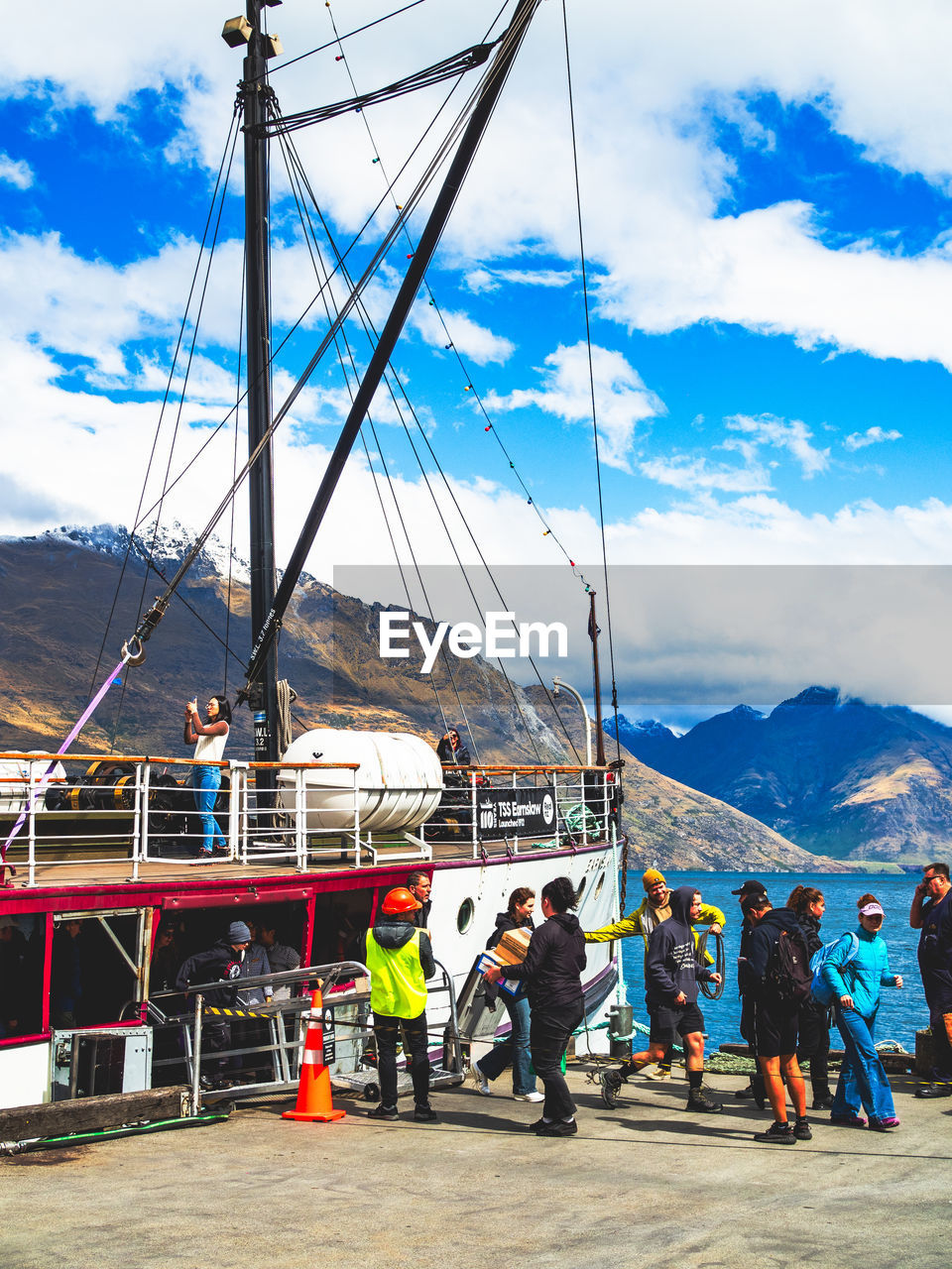 sky, cloud, transportation, nautical vessel, vehicle, group of people, water, nature, mountain, sea, men, mode of transportation, ship, travel, day, sailboat, adult, occupation, boat, outdoors, architecture, harbor, mast, pole, standing, pier, travel destinations, watercraft, sailing