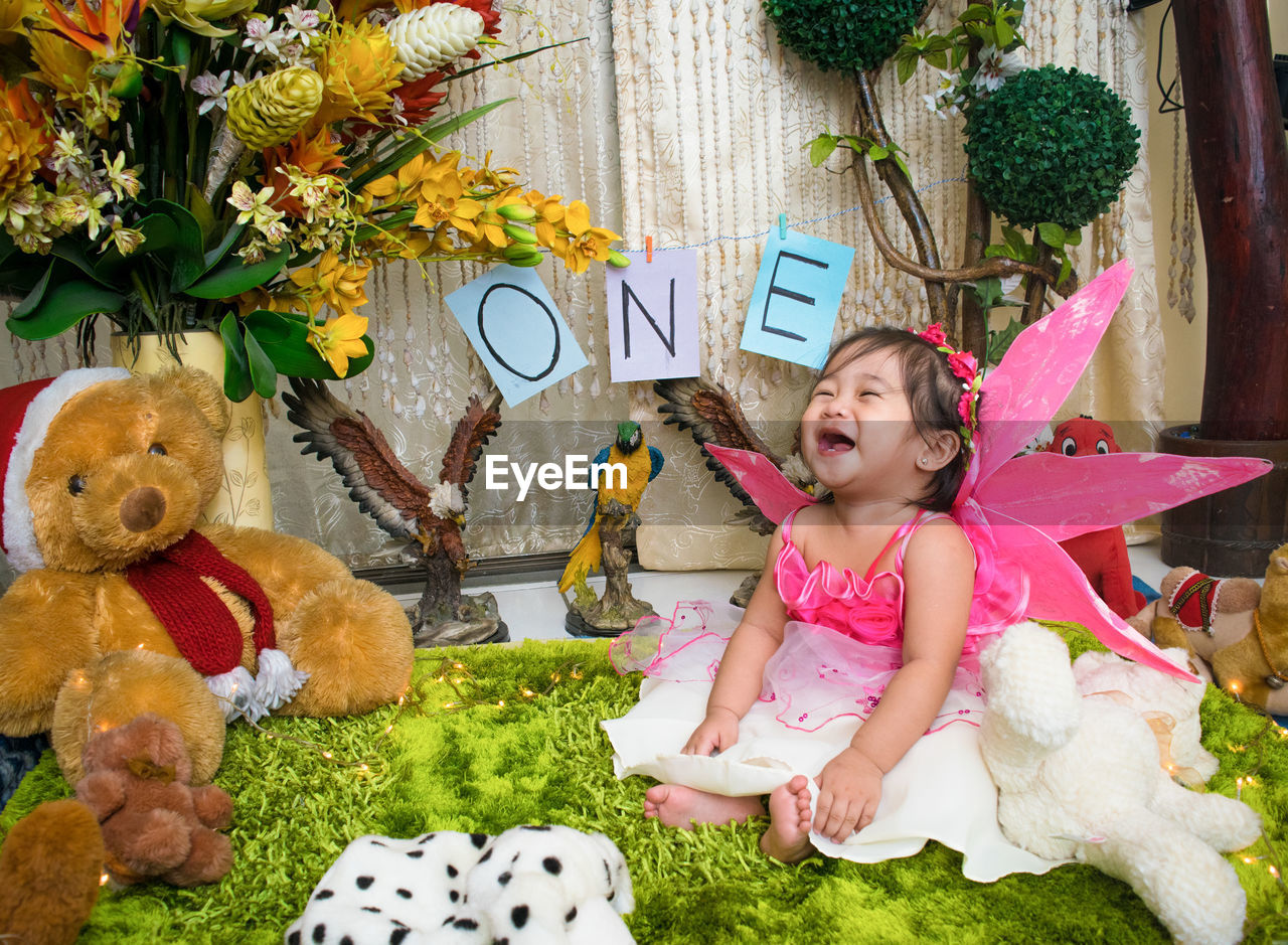 Cheerful shirtless baby girl sitting amidst stuffed toys during birthday