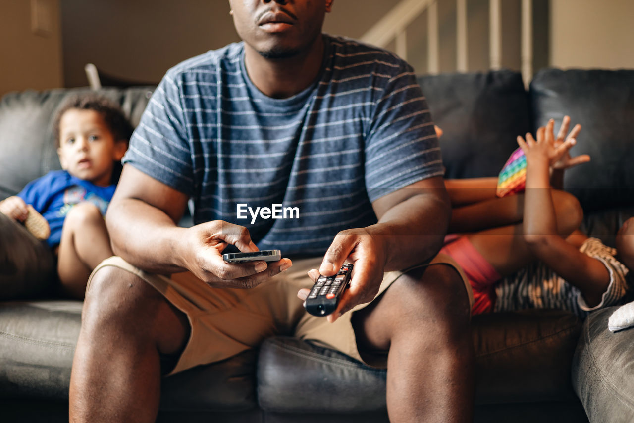 Father at home sitting on couch with kids in the background while holding tv remote and cellphone