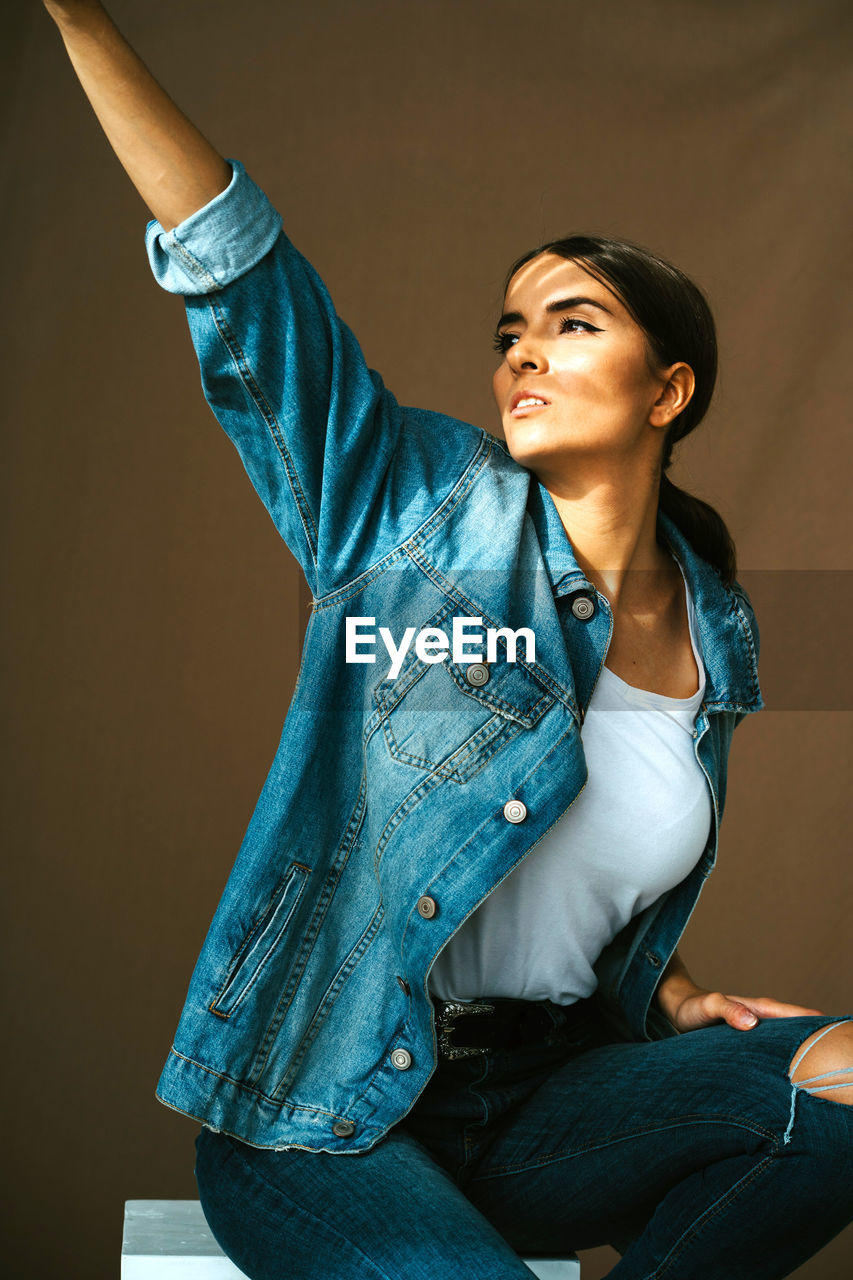 Confident young brunette in denim jacket raising hand and covering eyes from bright sunlight while standing against beige wall looking away