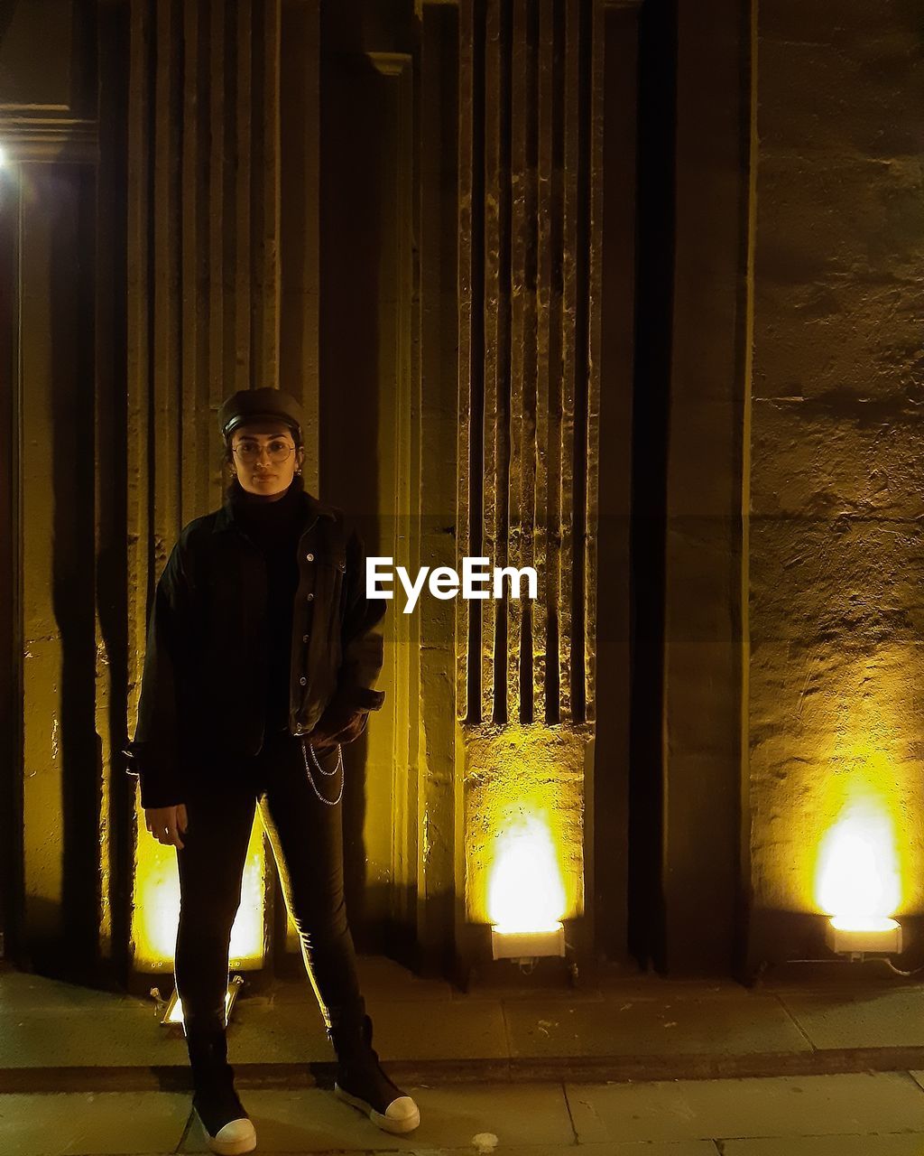 Portrait of woman standing on footpath by illuminated lights at night