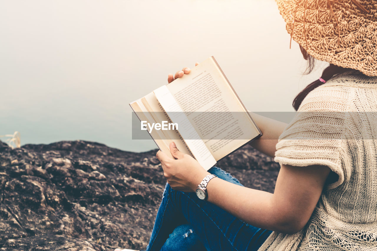 Midsection of woman reading book while sitting on rock by lake