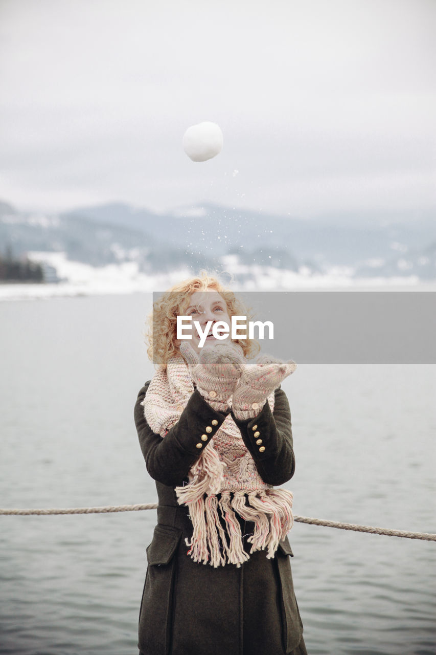 Happy woman playing with snowball by lake against sky