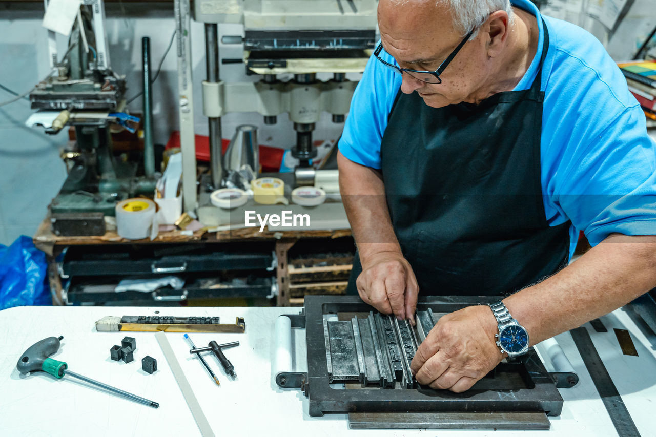 High angle of crop serious elderly male artisan in apron and eyeglasses preparing metal frame for letterpress printing in studio