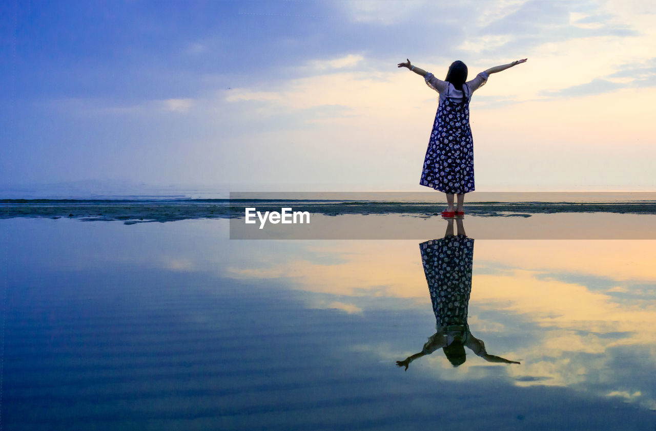 Carefree woman standing at seashore against sky during sunset