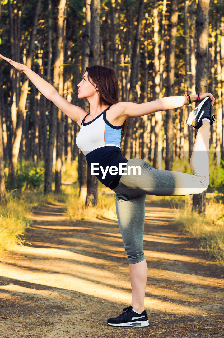 Full length of young woman exercising while standing against trees in forest