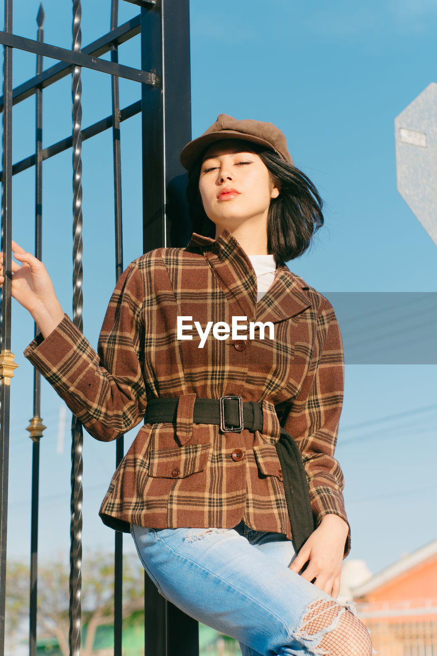 From below young ethnic female in checkered jacket with red lips looking at camera against fence in daytime