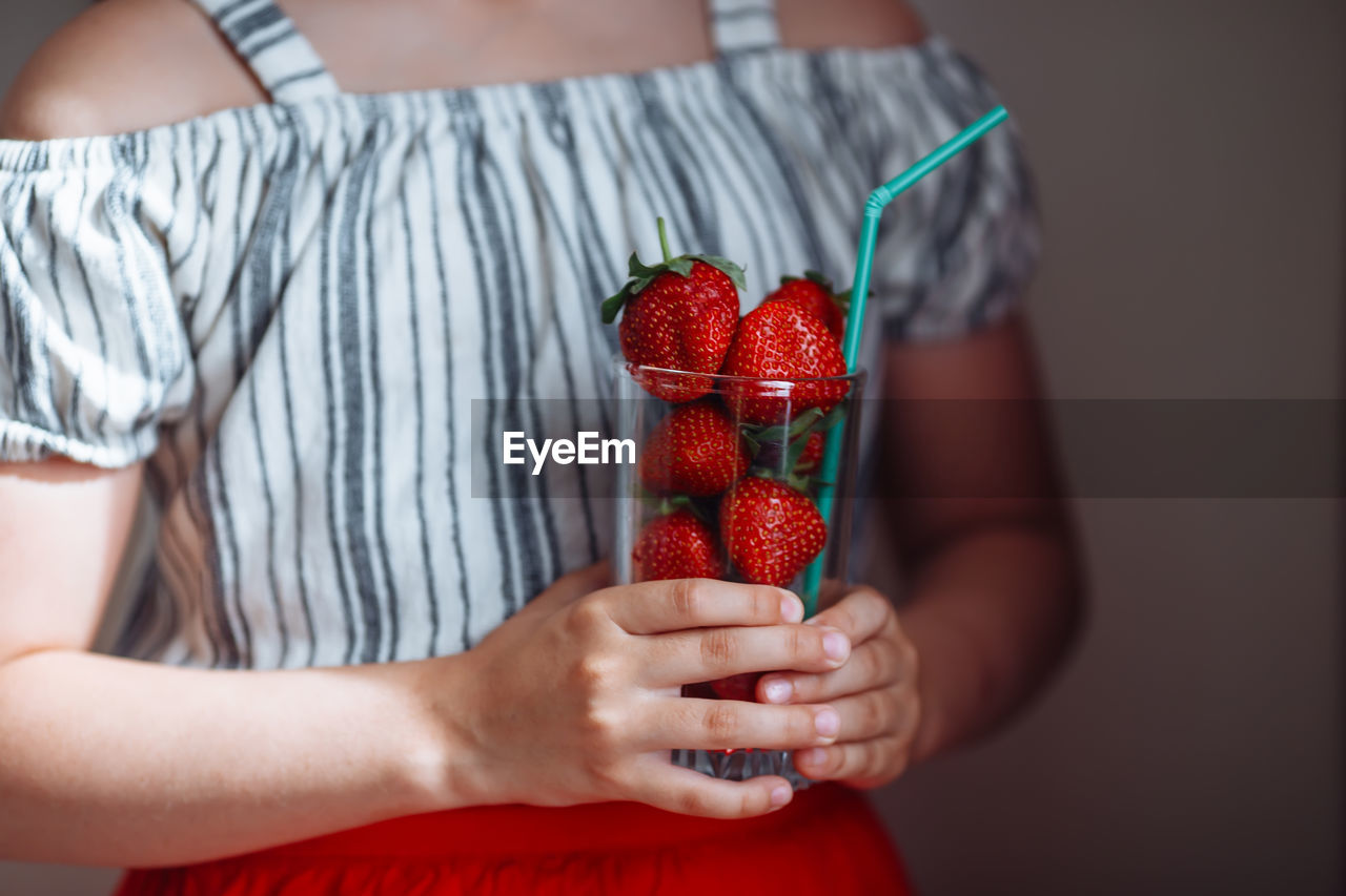 The hands of a girl in a striped blouse and a red skirt hold a glass with juicy strawberries.