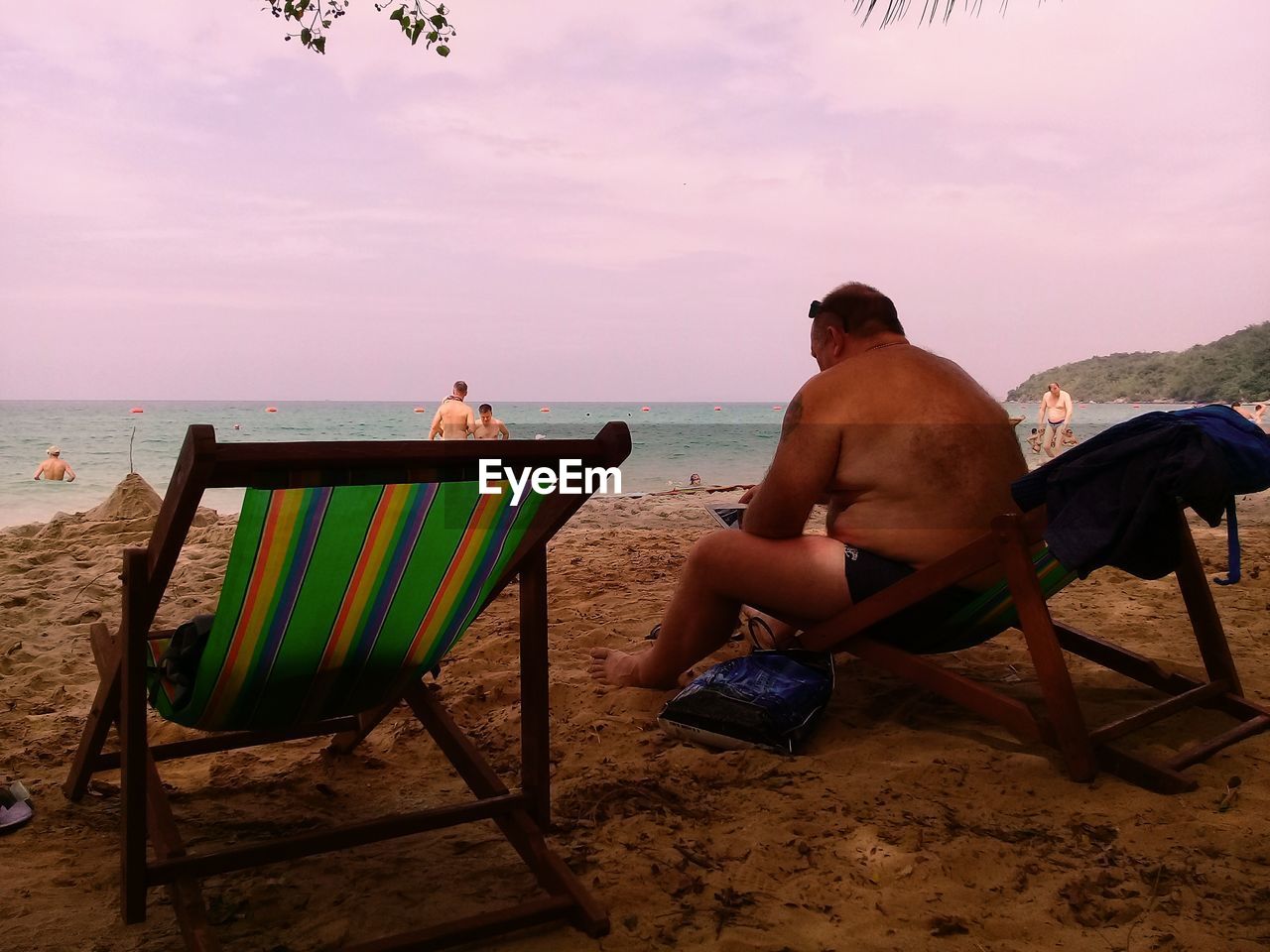 REAR VIEW OF SHIRTLESS MAN SITTING ON CHAIR AT BEACH AGAINST SKY