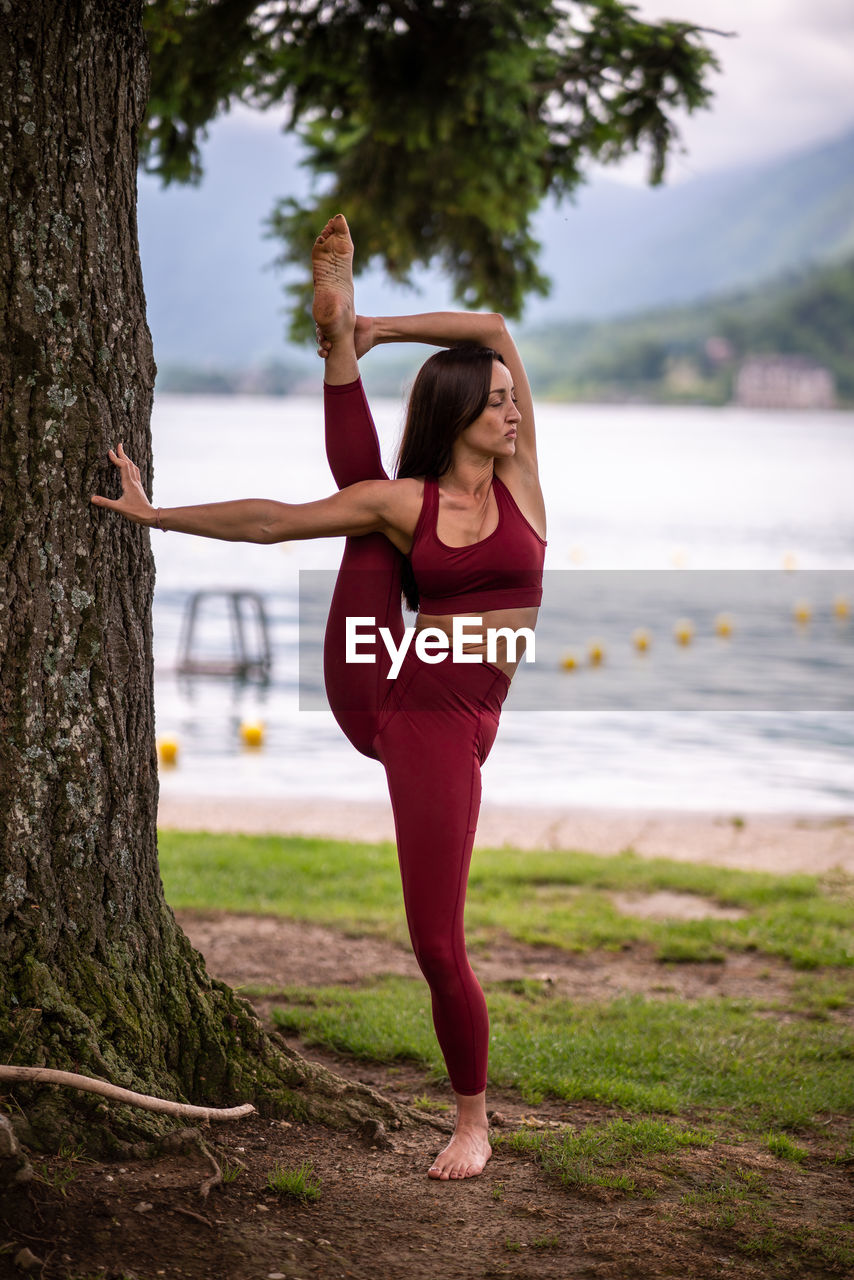 Slim female standing in svarga dvijasana while doing yoga on shore of pond and leaning on tree