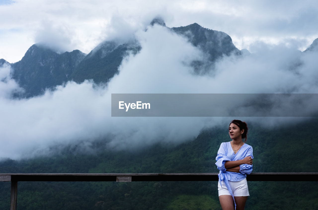 Beautiful woman standing by railing against mountains amidst fog