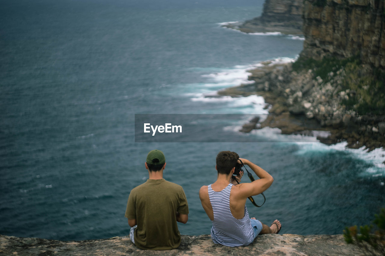 Rear view of male friends sitting on cliff against sea