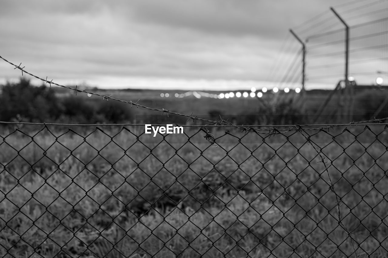 fence, chainlink fence, sky, security, cloud, wire, protection, monochrome, black and white, wire fencing, black, nature, no people, monochrome photography, barbed wire, focus on foreground, metal, line, wire mesh, outdoors, day, environment, architecture, outdoor structure, chain-link fencing, land