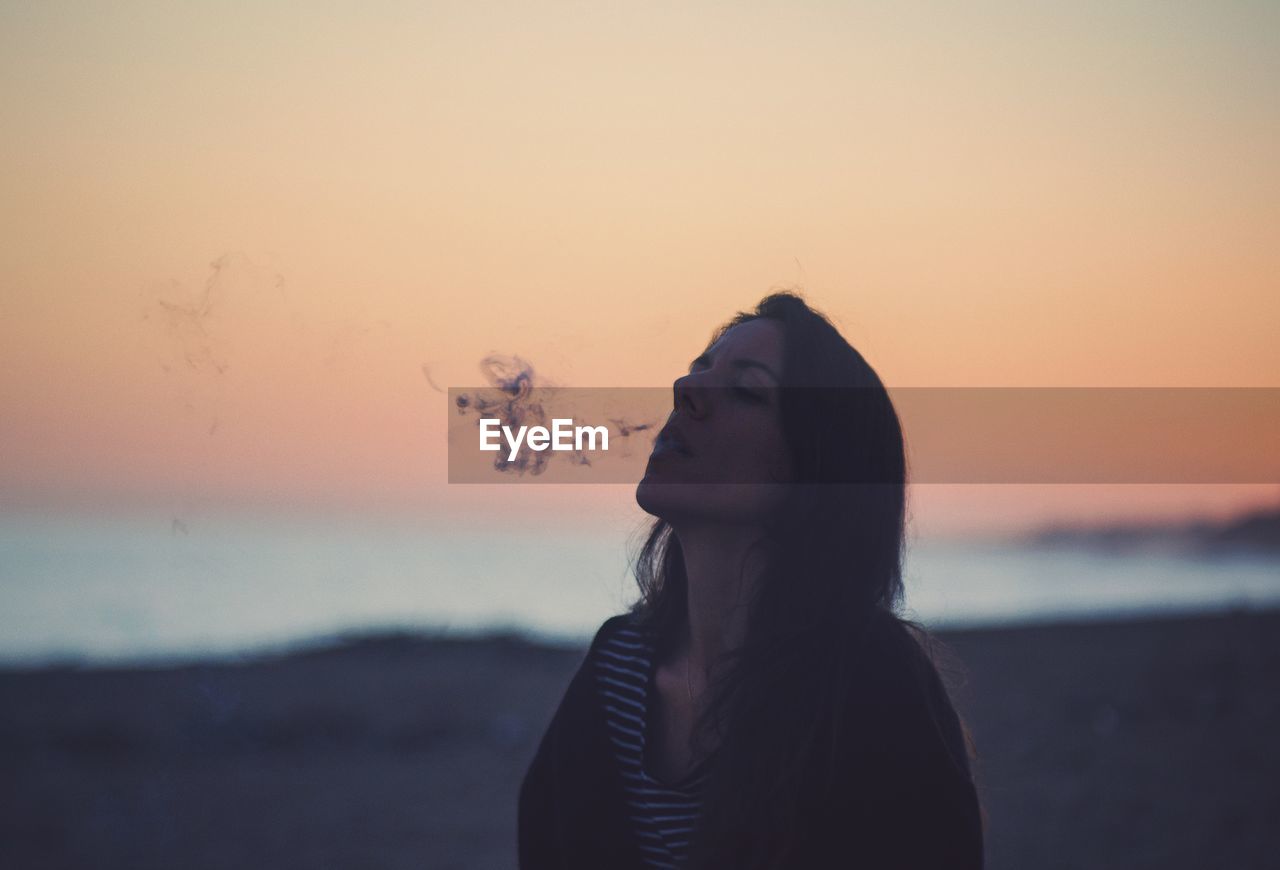 Mature woman smoking while standing at beach against sky during sunset