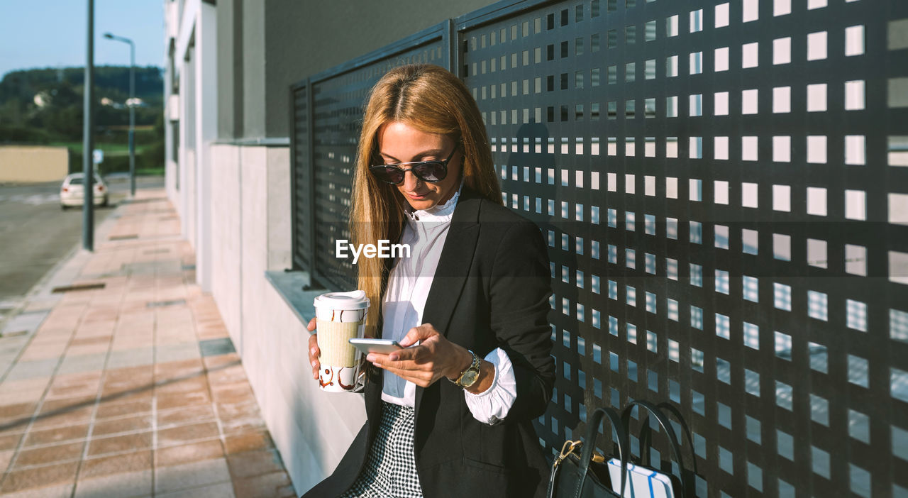 Businesswoman using smart phone holding coffee cup outdoors