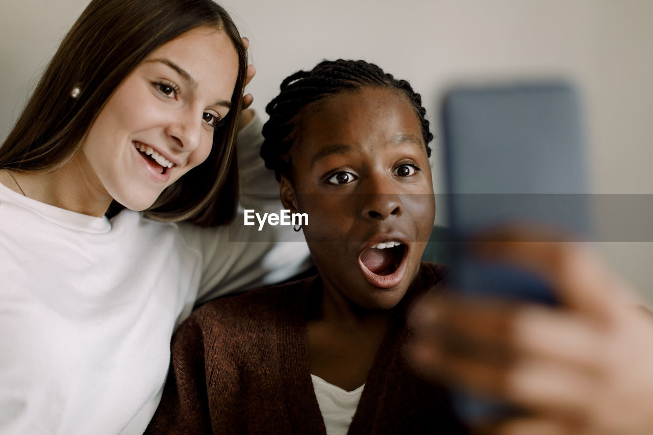 Shocked teenage girl looking at smart phone while sitting by smiling friend selfie at home