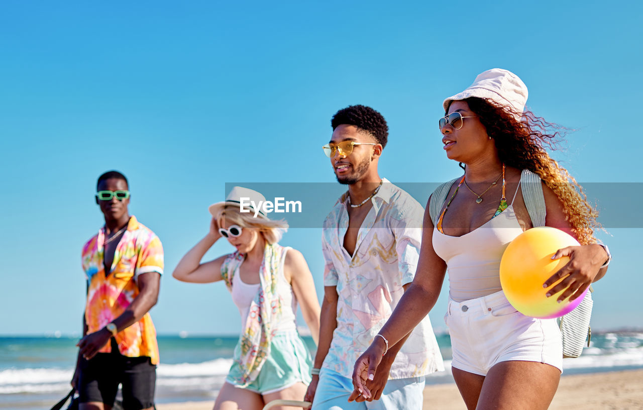Full body smiling multiracial men and women in sunglasses and hat strolling on sand against cloudless blue sky while spending time on beach on summer day