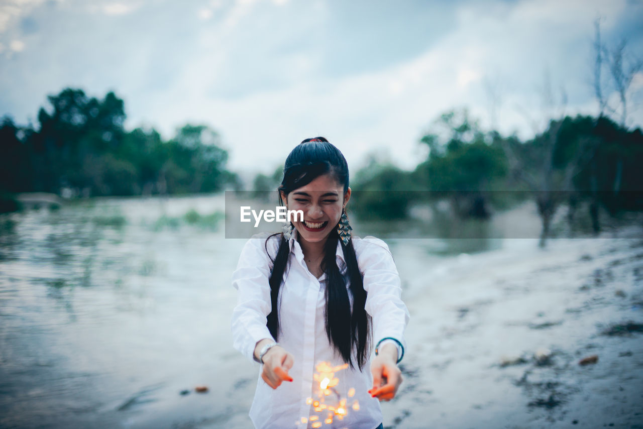 Cheerful woman holding lit sparklers at lakeshore