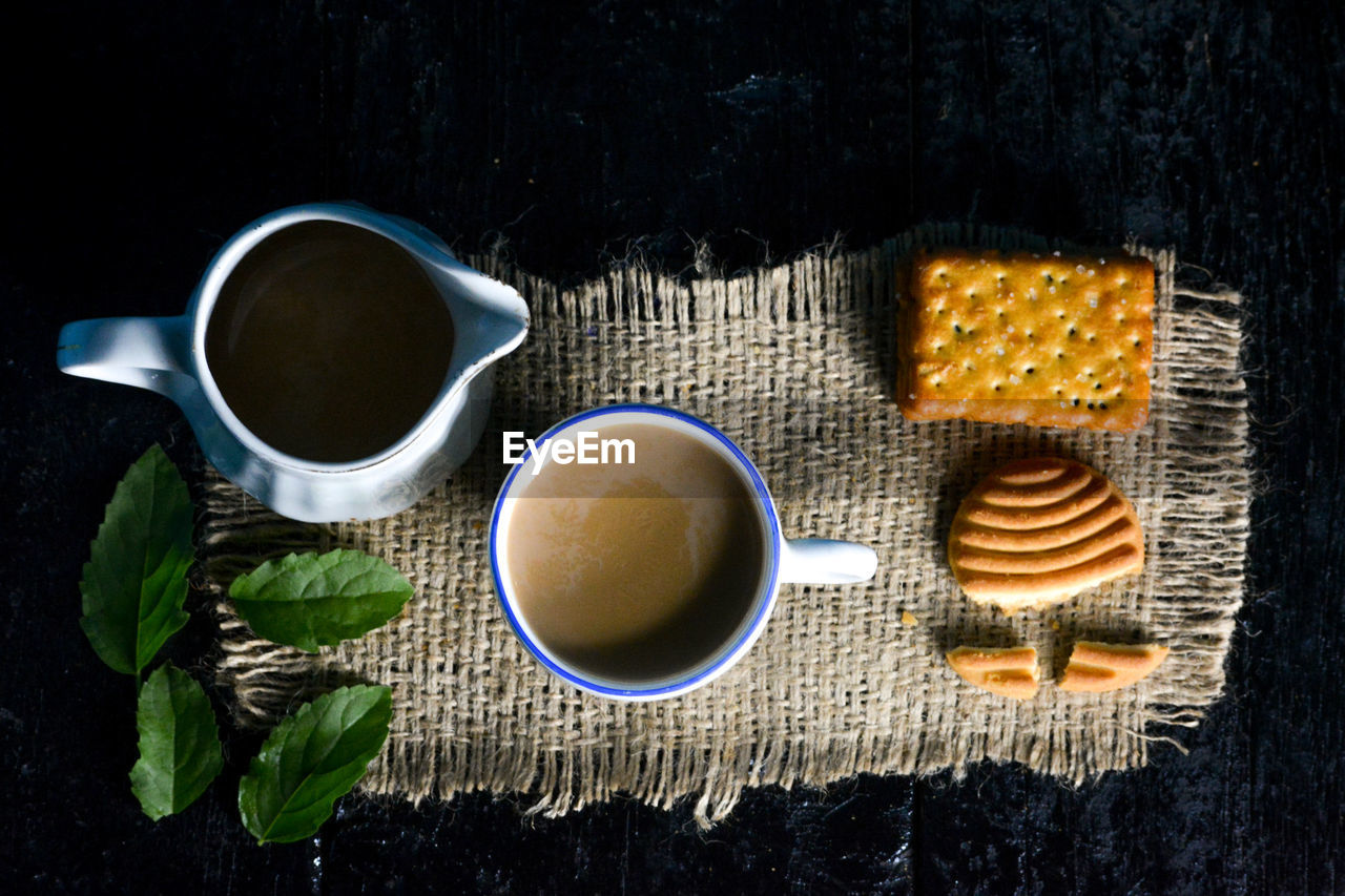 A cup of tea with tasty biscuits, teapot and fresh leaves on old wooden dark background