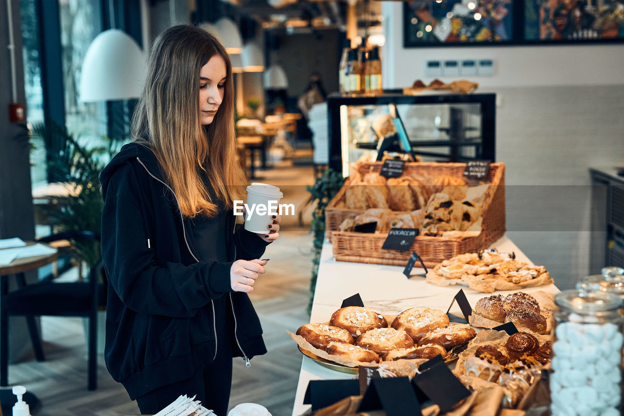 Woman holding cup with coffee looking at pastry, buns, cakes and cookies and waiting for the order