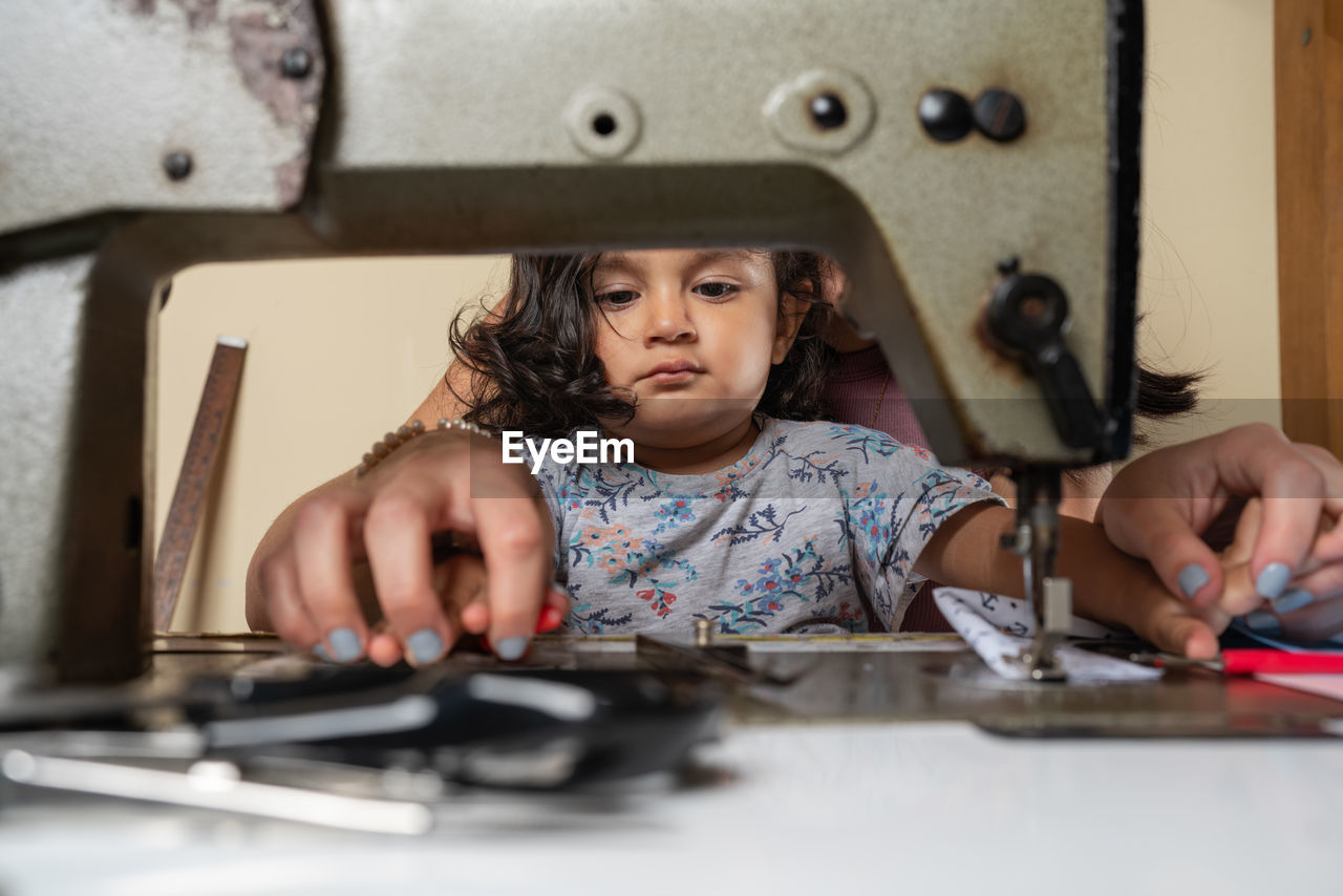 Unrecognizable seamstress with cute hispanic girl sewing on professional rusty machine while sitting at table with supply in light workshop