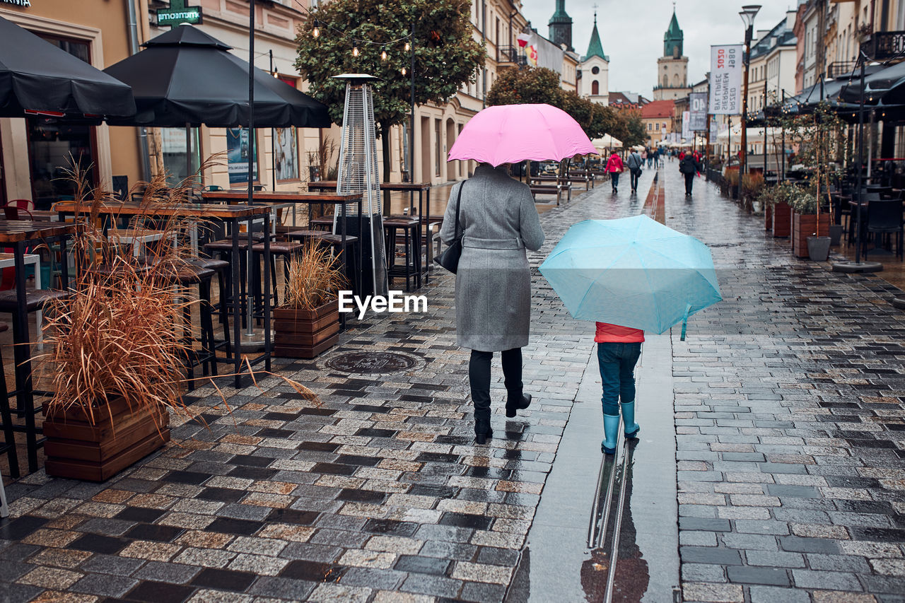 Back view of mother and her daughter holding the pink and blue umbrellas walking in a downtown