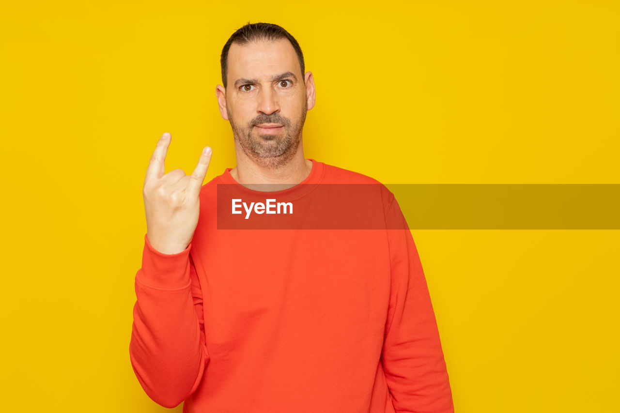 yellow, one person, studio shot, adult, portrait, colored background, looking at camera, waist up, men, finger, indoors, gesturing, yellow background, beard, facial hair, casual clothing, emotion, front view, sign language, person, orange color, young adult, clothing, hand, smiling, copy space, hand sign, human face, standing, orange, red, thumbs up, happiness, individuality, communication, vibrant color, positive emotion, t-shirt