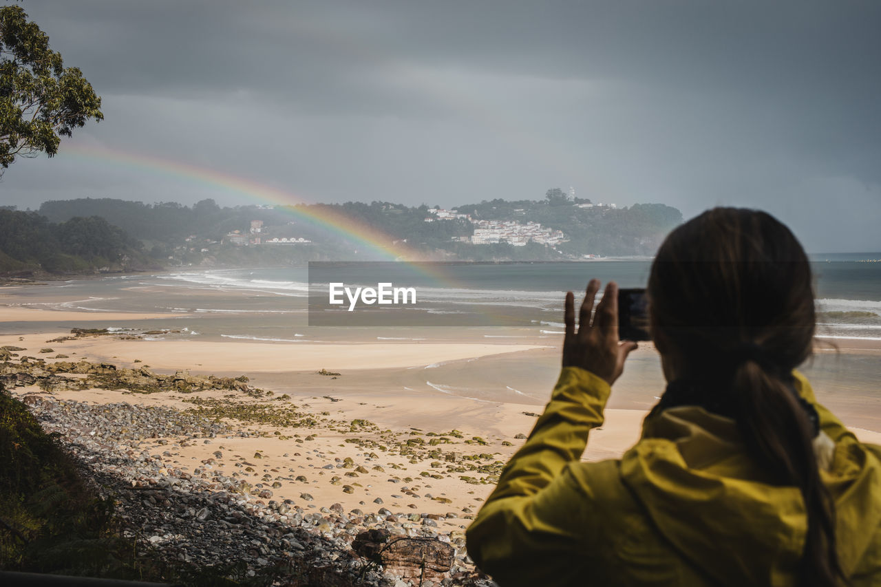 Rear view of woman in raincoat photographing rainbow at beach