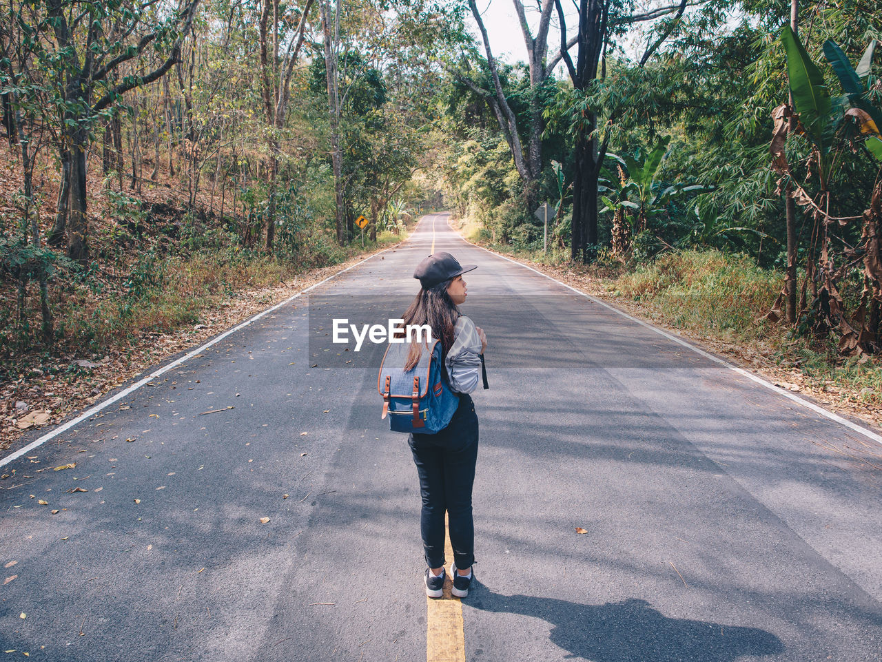 Woman hiking with backpack walking on roadway amid greenery trees.