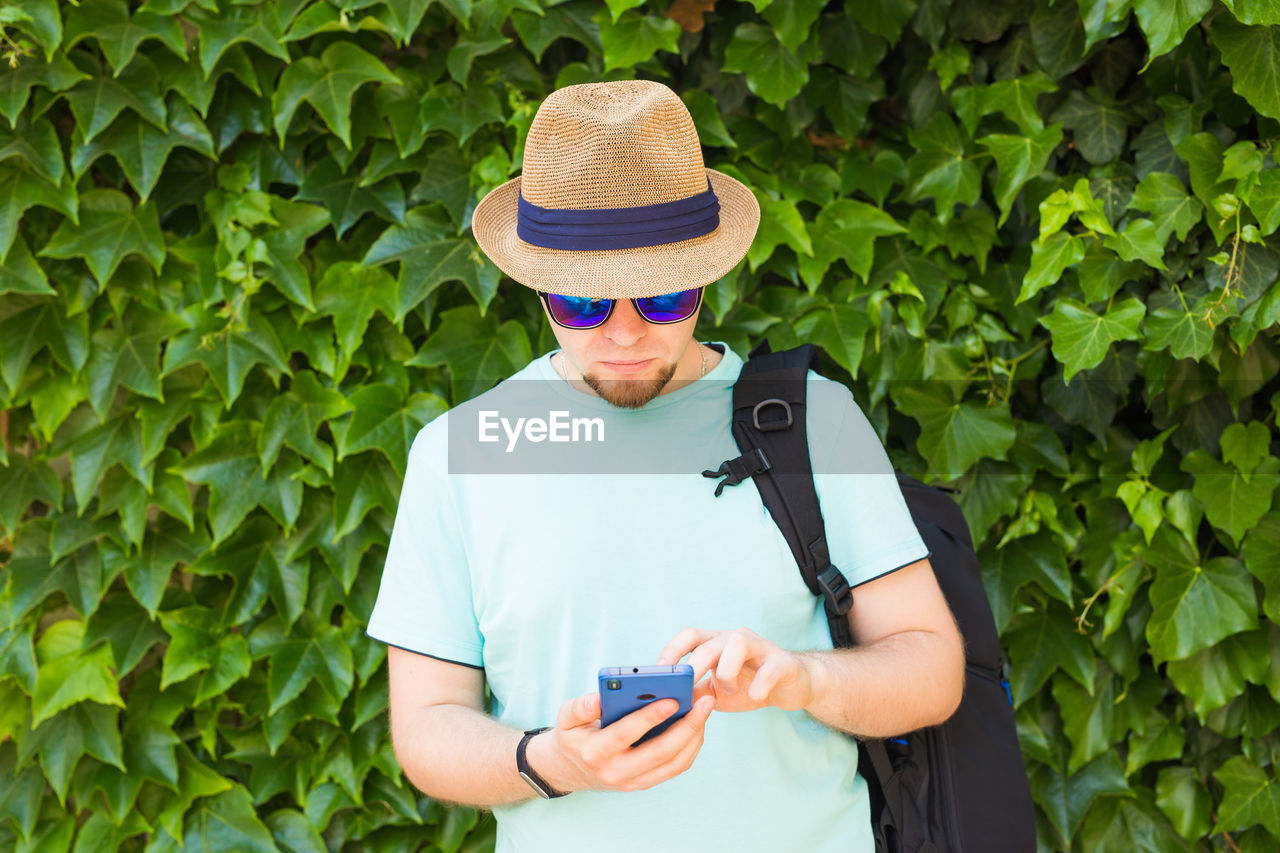 MIDSECTION OF MAN USING MOBILE PHONE WHILE STANDING BY PLANTS