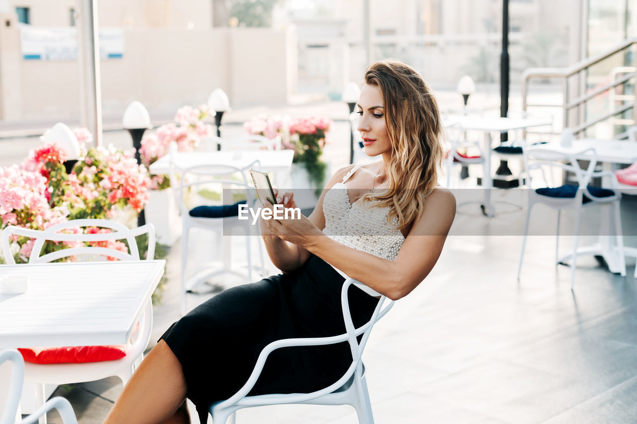 A young stylish woman sits in a street cafe and uses her phone to exchange messages 