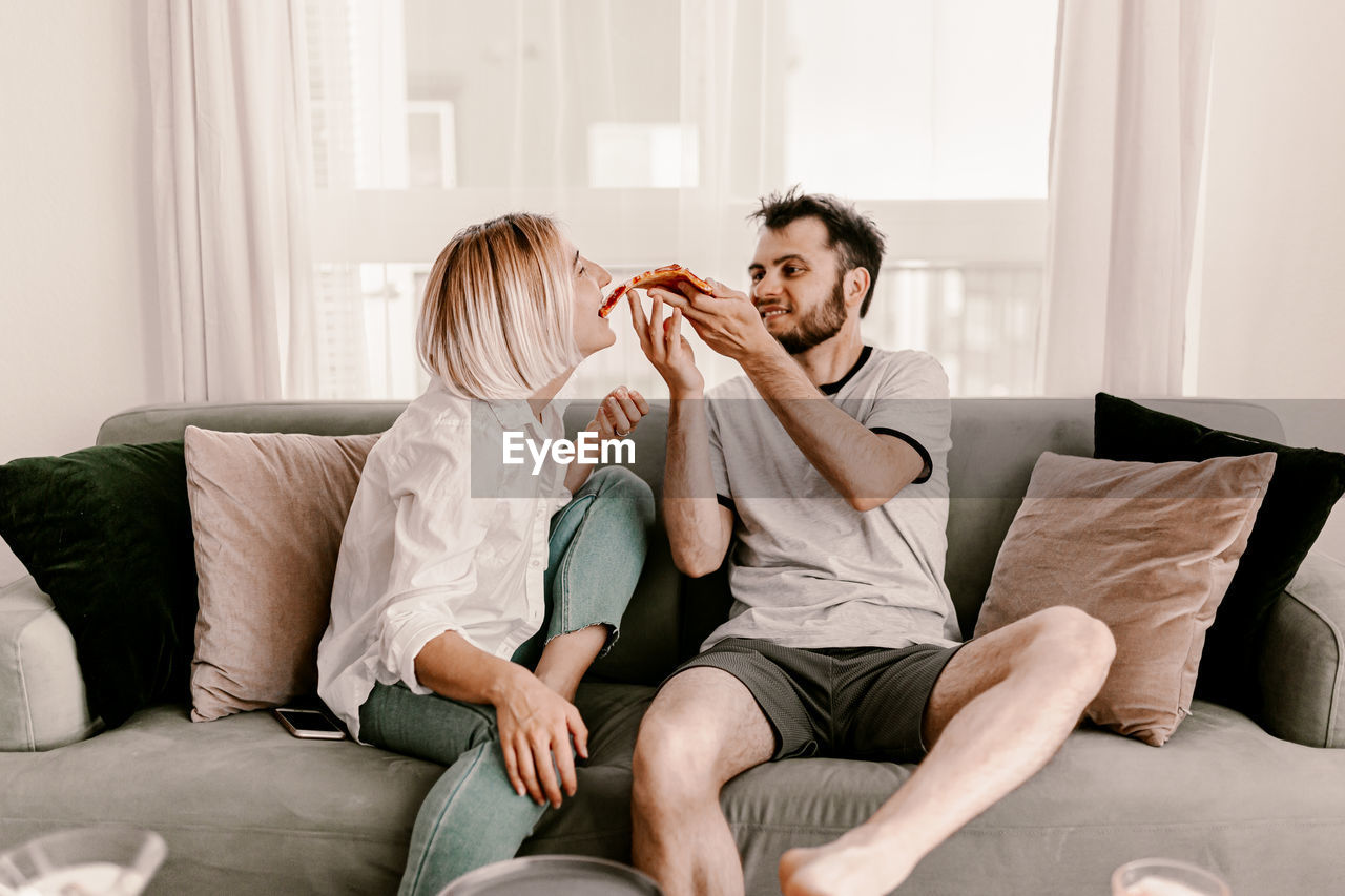 Man feeding pizza to girlfriend while sitting on sofa at home