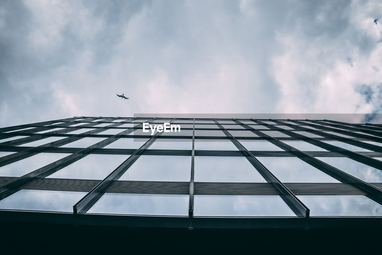 Low angle view of bird flying over building against cloudy sky