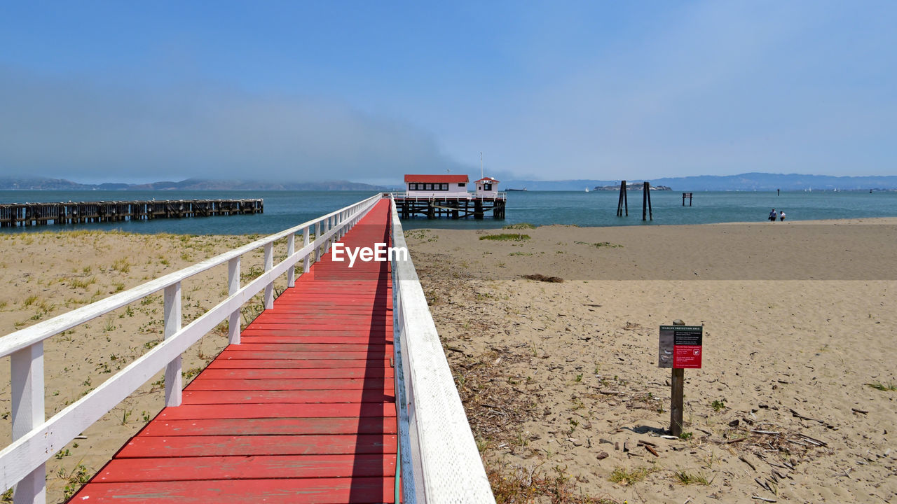 beach, land, sea, sky, water, walkway, shore, body of water, coast, sand, nature, boardwalk, scenics - nature, ocean, beauty in nature, horizon over water, vacation, horizon, travel destinations, tranquility, red, tranquil scene, travel, architecture, pier, no people, holiday, day, clear sky, tourism, transportation, blue, trip, man made structure, outdoors, coastline, wood, non-urban scene, landscape, lifeguard, cloud, environment, sunny, summer, built structure, lifeguard hut, railing, hut, sunlight, water's edge, idyllic, the way forward, bay, seascape, absence