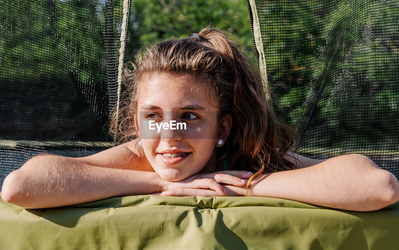 Horizontal portrait of a curly brunette teenager very happy smiling while lying on a trampoline with the net at the background. the girl feels happy and tired after jumping on the trampoline.