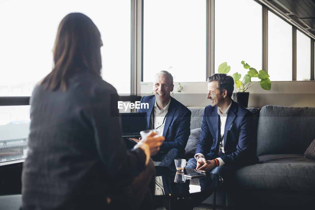 Smiling businessmen with laptop looking at female colleague while sitting on sofa in office