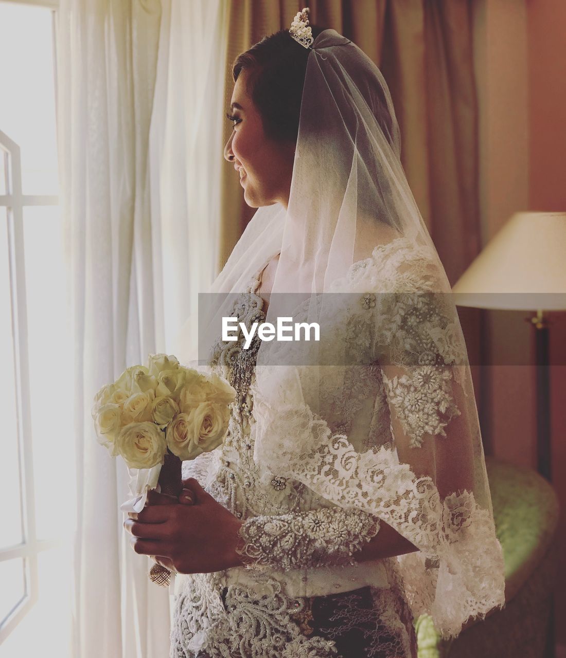 Smiling bride looking away while holding flower bouquet by window