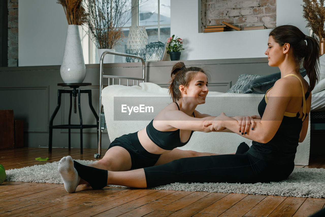 Positive athletic women with neatly tied hair and tight sportswear stretching at home. 