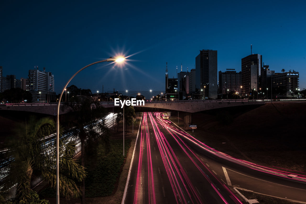 LIGHT TRAILS ON ROAD AMIDST ILLUMINATED BUILDINGS IN CITY AT NIGHT