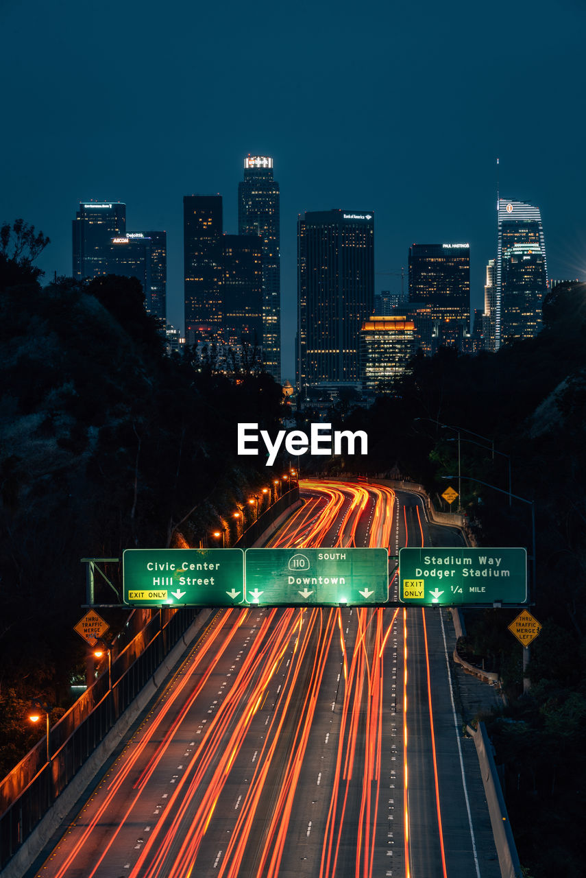 LIGHT TRAILS ON ROAD AMIDST BUILDINGS AGAINST SKY AT NIGHT