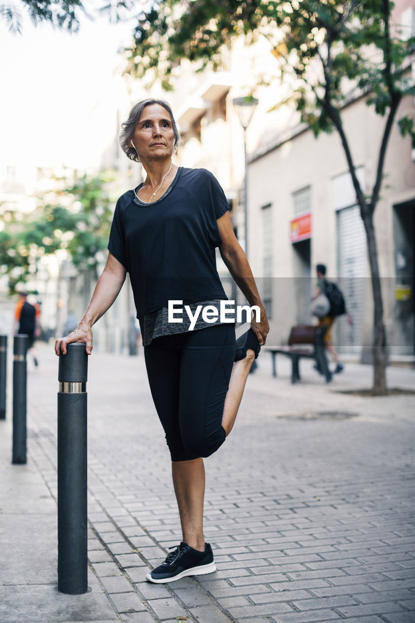 Low angle view of woman stretching leg while standing by bollard on footpath