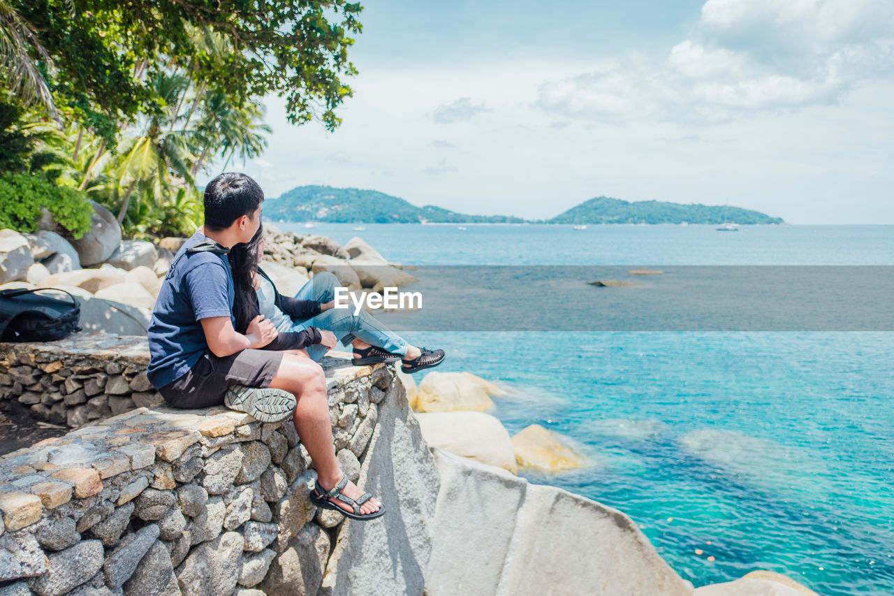 Rear view of woman sitting on rock by sea against sky