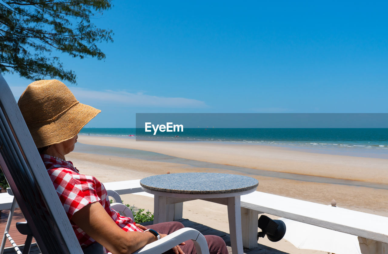 Senior woman sitting on balcony looking at blue sea and white beach in sunny day of summer vacation