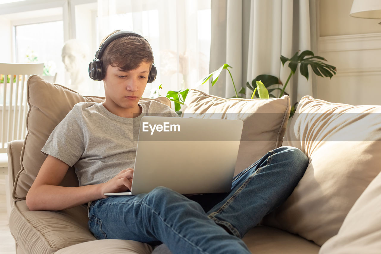 Portrait of teenager boy lying on a sofa in a room, with black headphones and laptop