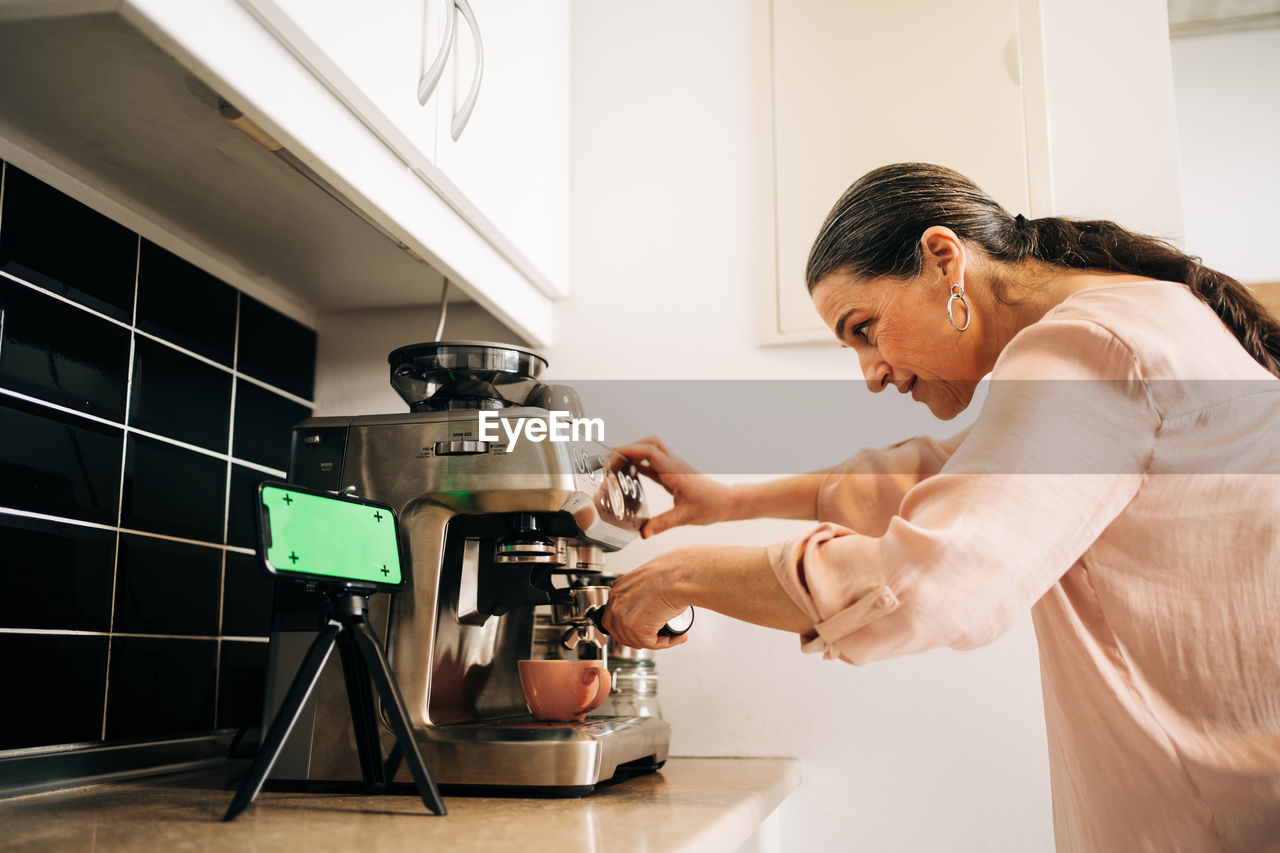 Side view of concentrated middle aged female preparing fresh coffee while using modern coffee maker at kitchen counter with smartphone on tripod