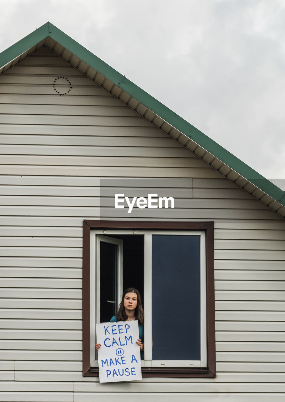 Teen girl in the window of her house. she is holding a poster. keep calm make pause